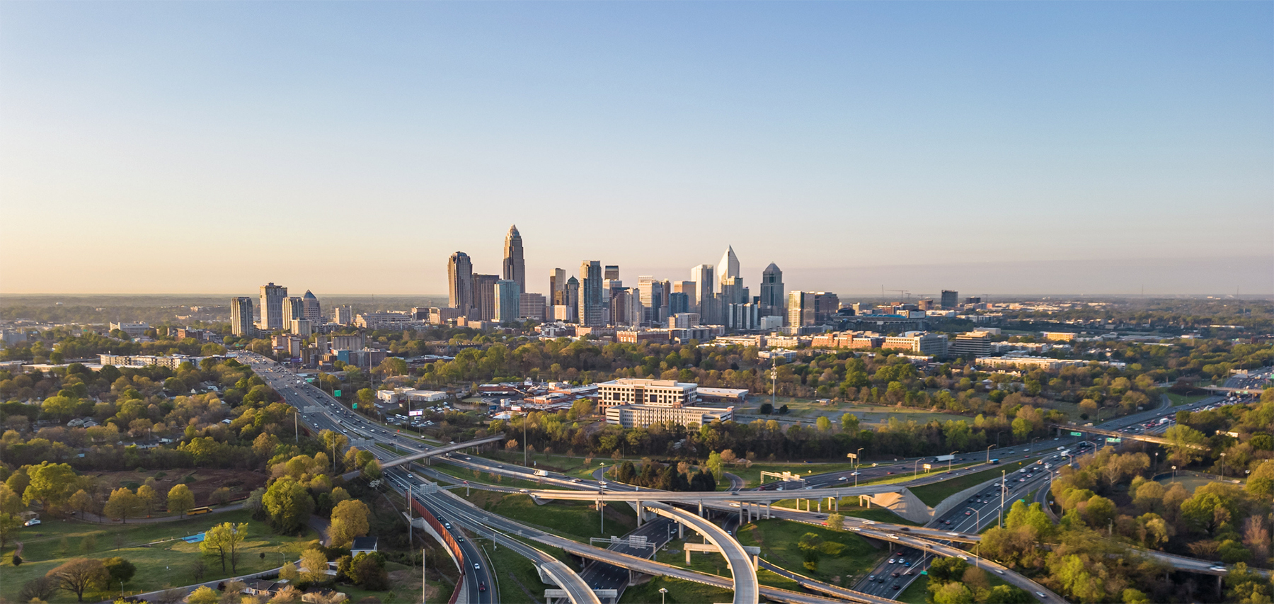 Aerial view of downtown Charlotte, North Carolina, with modern skyscrapers, highways, and surrounding greenery at sunset.