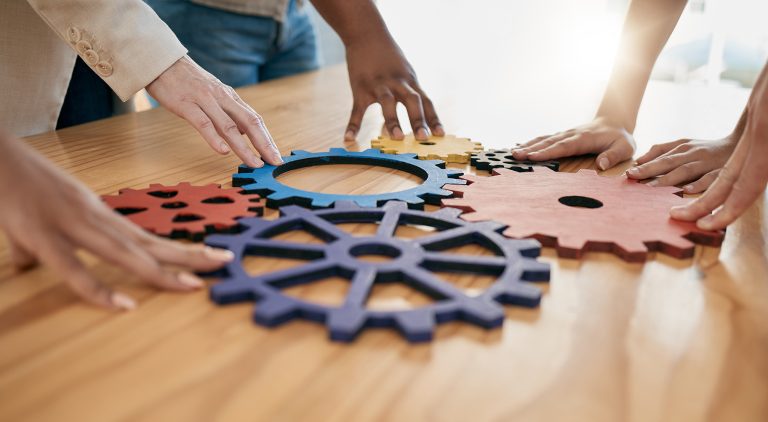Hands of diverse people connecting colorful gear-shaped pieces on a table.