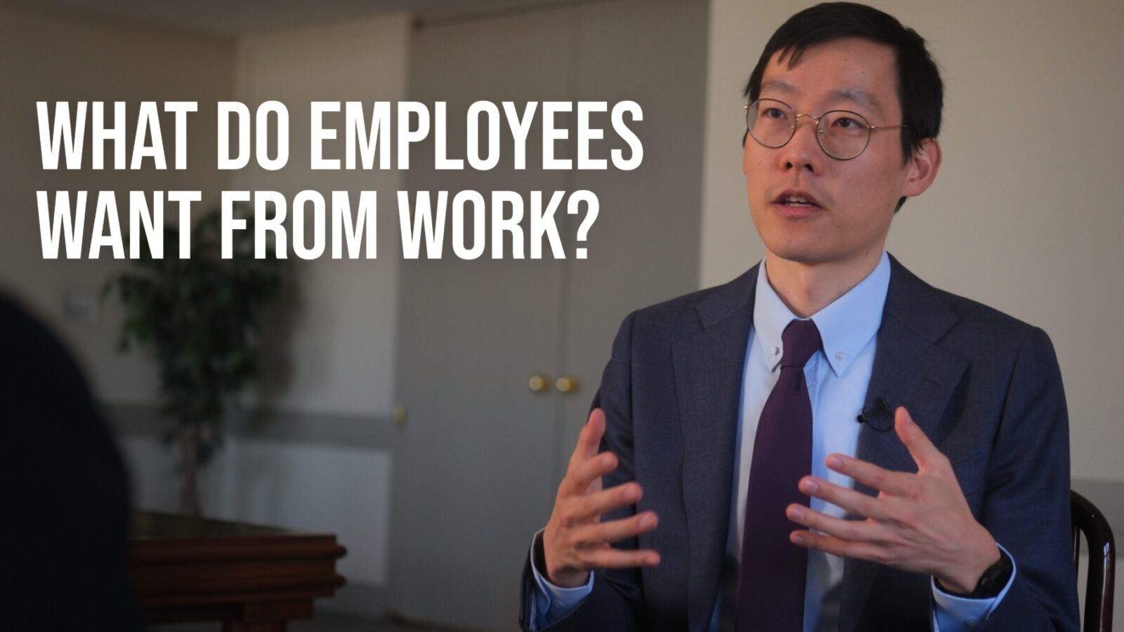 Professional speaking scene with a person in a suit and glasses gesturing while seated indoors. Large overlay text reads: “WHAT DO EMPLOYEES WANT FROM WORK?”