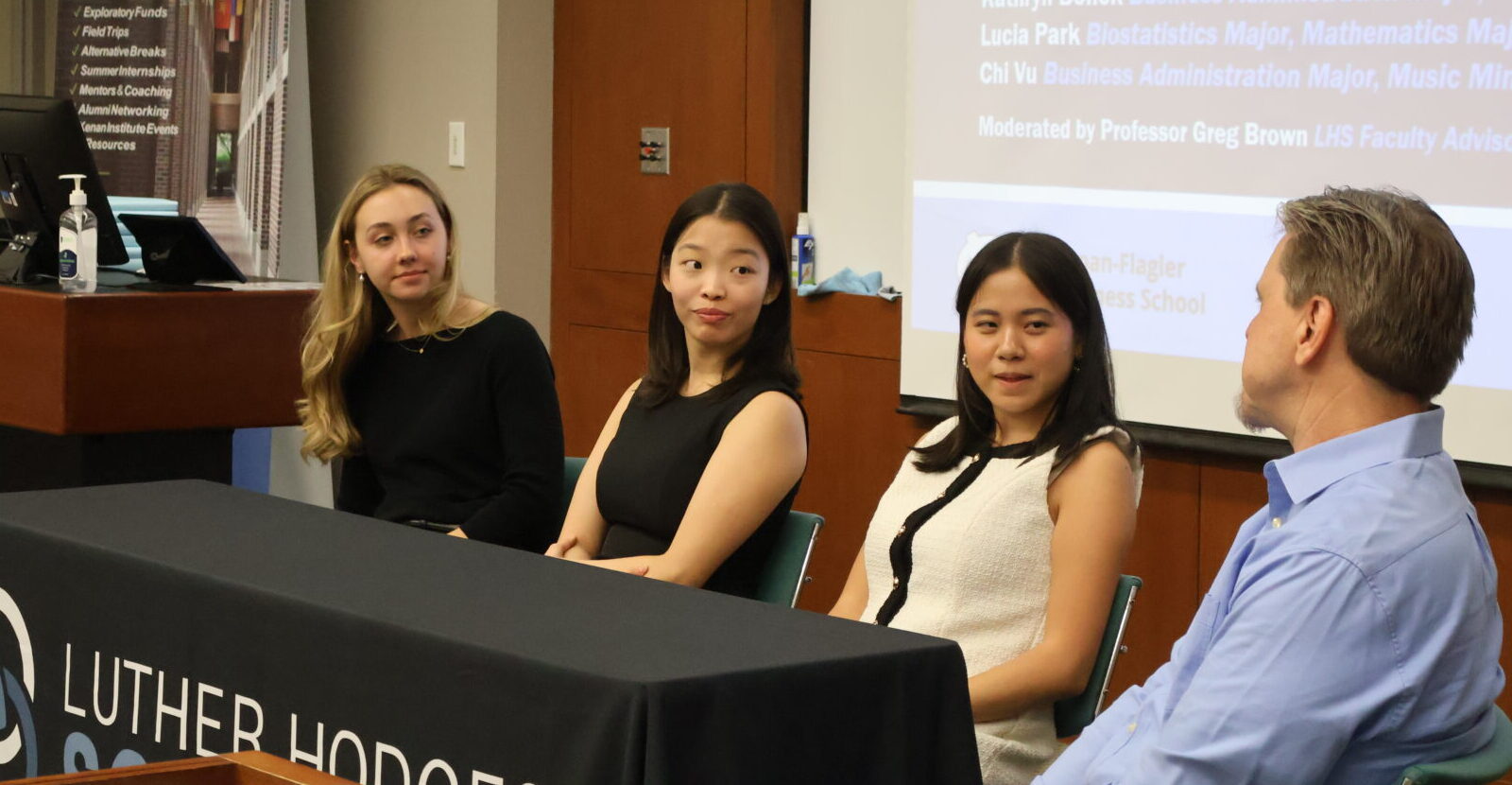 Three woman and a man sitting on a panel at a conference in front of a powerpoint presentation