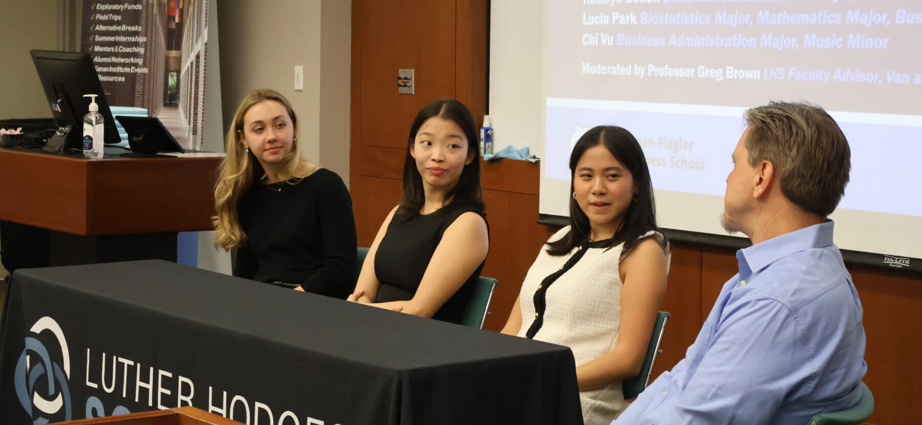 Three woman and a man sitting on a panel at a conference in front of a powerpoint presentation