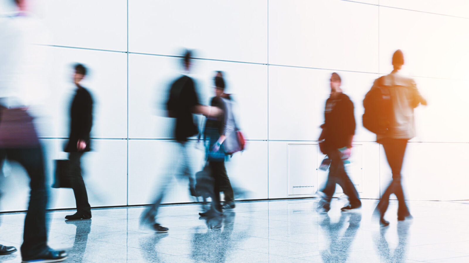 Motion-blurred pedestrians walk through a bright, modern indoor corridor with a glossy reflective floor and large white wall panels. Several people carry bags or backpacks.