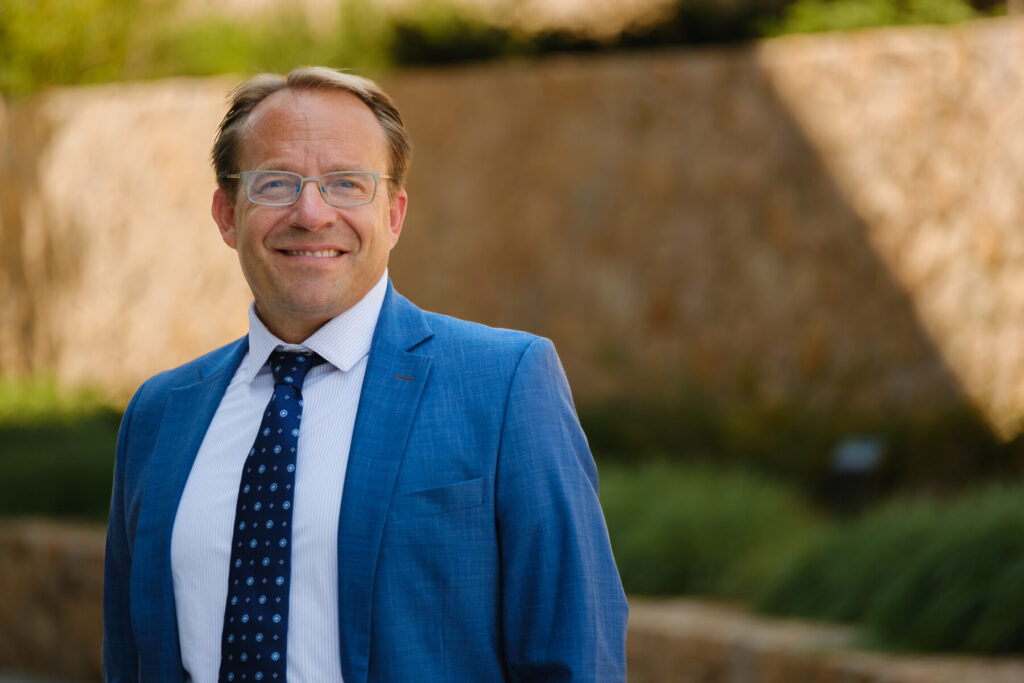 Professional outdoor headshot of a person wearing glasses, a blue suit jacket, white dress shirt, and patterned blue tie. The person stands in the foreground with a blurred background of stone walls and green landscaping in natural daylight.