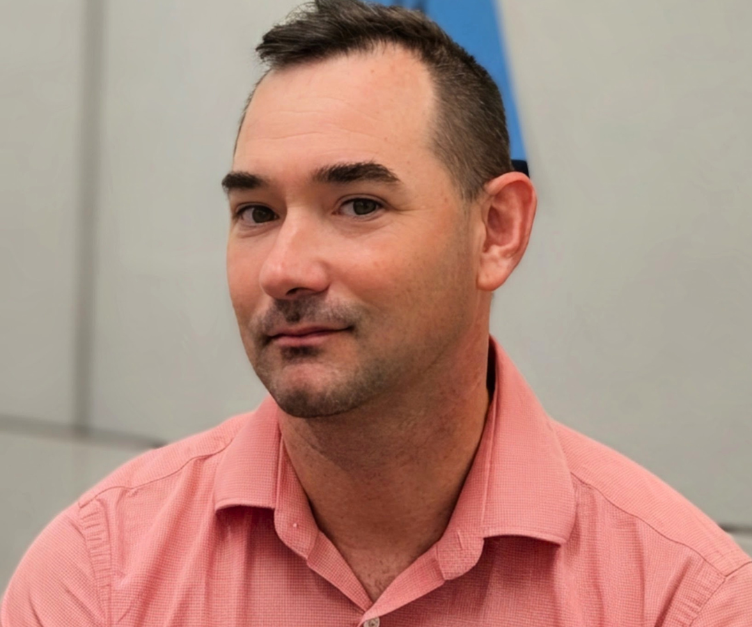 Portrait of Headshot of Daniel Green in a button down shirt in front of a white wall.