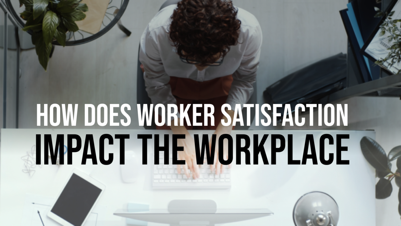 Overhead view of a person typing at a desk with a keyboard, tablet, and plants. Large text overlay reads: “HOW DOES WORKER SATISFACTION IMPACT THE WORKPLACE.”