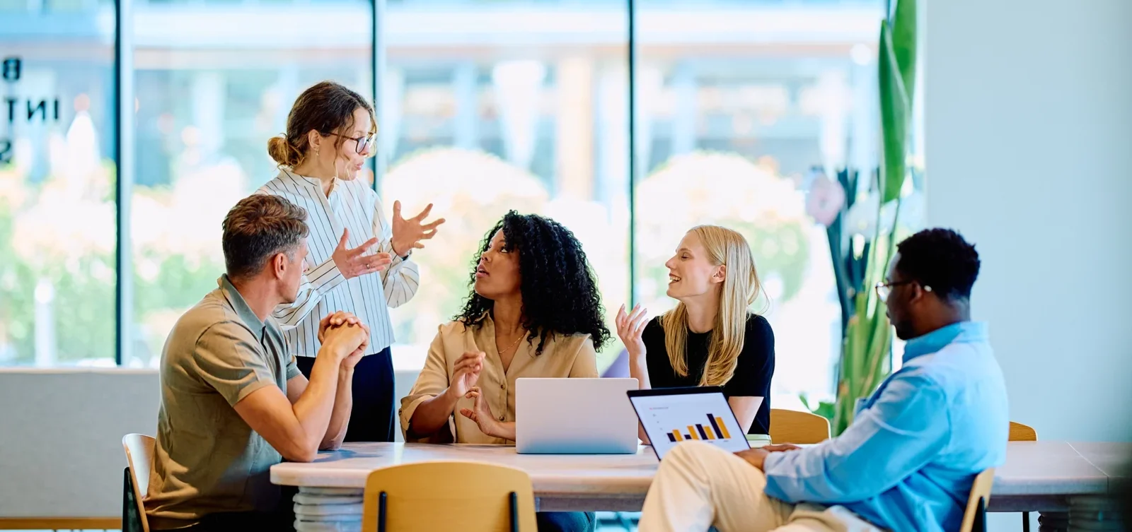 Five coworkers gather around a table in a bright, modern office, engaged in a lively discussion with laptops.