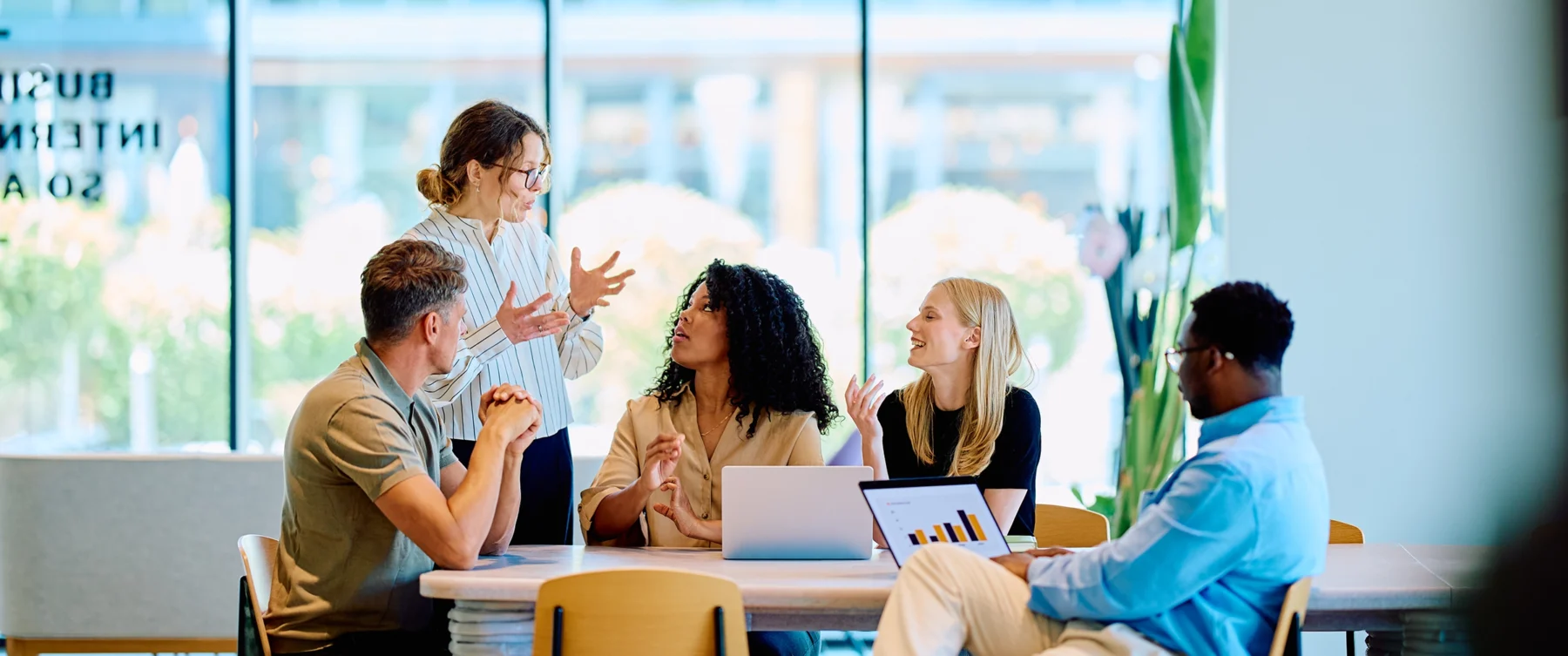 Five coworkers gather around a table in a bright, modern office, engaged in a lively discussion with laptops.